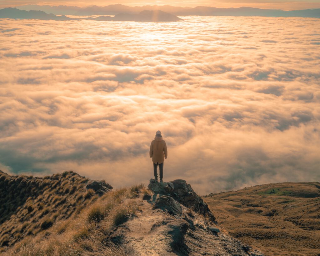 Person standing on top of mountain looking at sky filled with clouds and sunlight